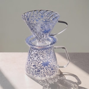 Op de afbeelding: Een koffiefilter en karaf van helder glas met een blauw bloemmotief van ranken. De conische filterhouder heeft interne ribbels en een handvat, terwijl de karaf een brede basis en een handvat van helder glas heeft.