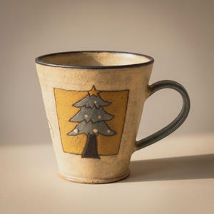 May include: A beige ceramic mug with a speckled texture and a dark rim. The front features a square panel containing a green pine tree illustration with a yellow star on top and small yellow dots, set against a mustard yellow background.