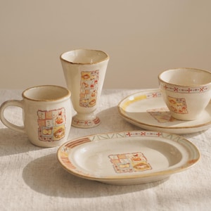 May include: A collection of cream-colored ceramic tableware featuring a repeating patchwork-style graphic print. The set includes a mug with a handle, a tall stemmed goblet, a small bowl on a matching saucer, and an oval-shaped serving plate.