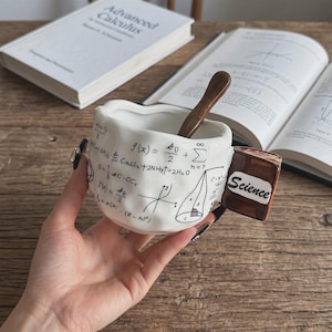 Peut inclure: Un mug en céramique blanche au bord et à la surface ondulés et irréguliers, orné de formules mathématiques et de diagrammes géométriques imprimés en noir. Il comprend une anse en forme de livre marron marquée Science et une cuillère en bois.