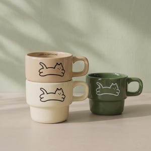 Può includere: Tre tazze in ceramica color beige, crema e verde oliva. Ogni tazza presenta un disegno a linee nere o bianche di un gatto in posizione sdraiata. La tazza beige superiore riporta la scritta I NEED A COFFEEINE stampata sul bordo interno.