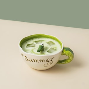 May include: A cream-colored ceramic mug with a green interior and a sculpted green handle. The exterior features the word Summer in brown script, a small sun icon, and a sleeping crocodile graphic. A 3D crocodile head emerges from the liquid.