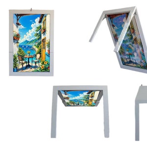 May include: A white picture frame with a hinged mechanism that allows the frame to fold down and become a table. The frame features a colorful painting of a seaside scene with a house, palm trees, and a blue sky.