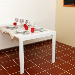 Könnte beinhalten: Ein weißer Klapptisch mit einer weißen Tischdecke und roten Gläsern. Der Tisch ist mit Tellern und Besteck gedeckt. Der Tisch steht in einem Raum mit Fliesenboden.