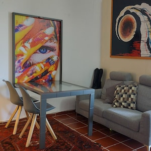 May include: A living room with a gray couch, two gray chairs, a glass-top table, and two framed paintings on the wall. One painting features a colorful abstract design, and the other features a black and white abstract design.