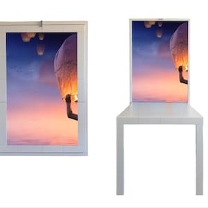 Könnte beinhalten: Ein weiß gerahmtes Bild mit einem Foto einer Hand, die eine brennende Himmelslaterne vor einem rosa und violetten Sonnenuntergangshimmel hält. Die Himmelslaterne schwebt in der Luft mit anderen Laternen im Hintergrund.