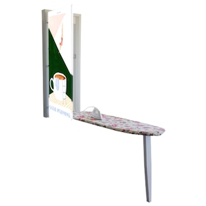 Op de afbeelding: Een wit strijkplank met een bloemenprint hoes is neergeklapt en bevestigd aan een wandmontage strijkplankhouder. De houder is bevestigd aan een witte deur met een groene en roze illustratie van een kopje koffie en een persoon die in een zwembad duikt. De illustratie bevat ook de tekst "Good Morning!"