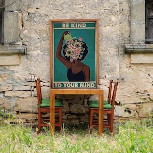Könnte beinhalten: Ein gerahmtes Poster mit dem Text "BE KIND TO YOUR MIND" zeigt eine Frau mit floralem Kopf und schwarzem Kleid. Das Poster wird auf einem Holztisch mit zwei Holzstühlen mit grünen Kissen ausgestellt. Der Hintergrund ist eine alte Steinmauer.