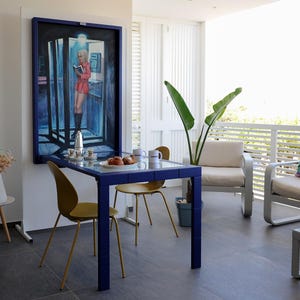 Könnte beinhalten: Ein blauer und weißer Esstisch mit vier Stühlen, ein weißes Sofa mit bunten Kissen und ein großes Gemälde einer Frau in einem roten Kleid, die in einer Telefonzelle steht. Der Tisch ist mit einem Teller Essen, einer Kaffeekanne und Tassen gedeckt.