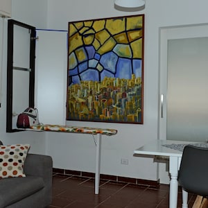 Puede incluir: Una tabla de planchar montada en la pared está abierta, con una funda con estampado floral. Una plancha roja y blanca está sobre la tabla. Una colorida pintura abstracta cuelga de la pared. Un sofá gris con un cojín de lunares está en primer plano.