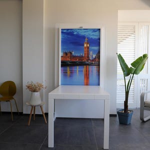 May include: A white table with a white framed picture of the London skyline at night. The picture includes Big Ben and the Houses of Parliament. The table is in a room with a white chair, a plant, and a white wall.