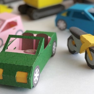 May include: A collection of colorful paper toy vehicles, including a green convertible, a pink car, a blue sedan, a yellow motorcycle, and a yellow construction vehicle. The vehicles are made of folded paper and have detailed features.