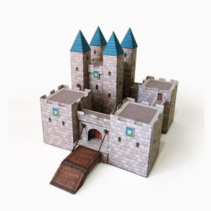 May include: A paper model castle featuring grey stone-textured walls, four tall towers with pointed blue roofs, and a brown drawbridge. The structure includes small arched windows, flat rooftops, and blue shields with yellow emblems on the exterior walls.