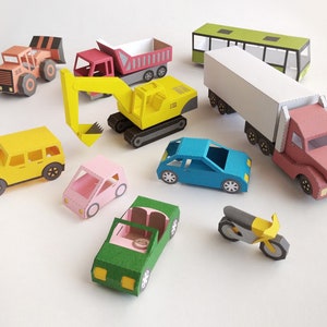 May include: A collection of colorful paper toy vehicles, including a yellow excavator, a red dump truck, a green bus, a semi-truck, and several cars. The toys are arranged on a white surface.
