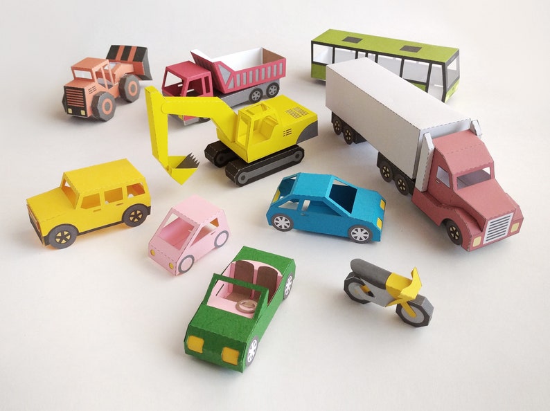 May include: A collection of colorful paper toy vehicles, including a yellow excavator, a red dump truck, a green bus, a semi-truck, and several cars. The toys are arranged on a white surface.