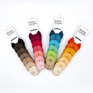 Könnte beinhalten: Vier Knäuel Braidy Ombre Garn in verschiedenen Farbverläufen. Das Garn ist ein Farbverlauf, wobei jedes Knäuel eine andere Start- und Endfarbe hat. Das Garn ist mit "Loopcraft Braidy Ombre yarn for knit" gekennzeichnet.
