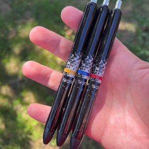 May include: Four black pens with silver tips and black rubber grips. Each pen has a white speckled design and a colored band: yellow, blue, and red. The pens are held in a hand against a blurred green background.