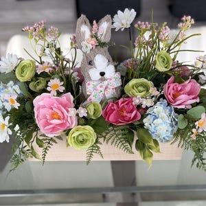 May include: A wooden rectangular planter box filled with colourful artificial flowers and greenery. A decorative bunny with a floral crown and bow tie is centered in the arrangement. The flowers include pink, white, green, and blue blooms.