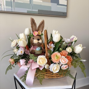 May include: A wicker basket filled with spring flowers and greenery, with a brown bunny figurine wearing a floral crown. The arrangement includes white tulips, peach roses, and a pink bow, creating a festive Easter centrepiece.