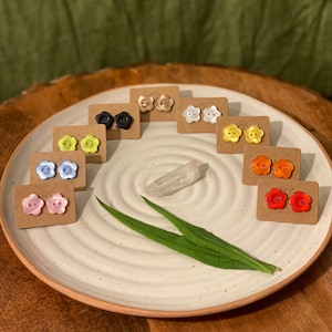 May include: Assortment of colorful flower-shaped button earrings displayed on a white plate with a green leaf. The earrings are arranged in a rainbow pattern, starting with gold, white, yellow, orange, red, pink, blue, black.