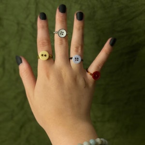 May include: A hand wearing four different button rings. The rings are made of gold, silver, and black wire. The buttons are white, yellow, and red.