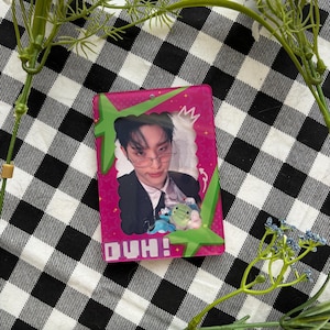 以下が含まれることがあります： メガネとスーツを着た人の写真が写ったピンク色のカードで、白い文字で「DUH！」と書かれています。カードは緑色のフレームと白い王冠で囲まれています。背景は白と黒のチェック柄で、白い花が飾られています。