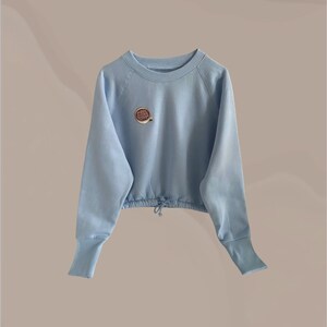 Peut inclure: Un sweat-shirt court bleu clair à manches longues et un cordon de serrage en bas. Le sweat-shirt a un petit motif de tasse à café brodé sur le devant.