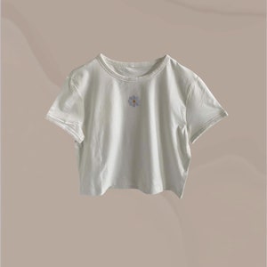 Peut inclure: Un t-shirt blanc court avec une petite fleur brodée au centre.