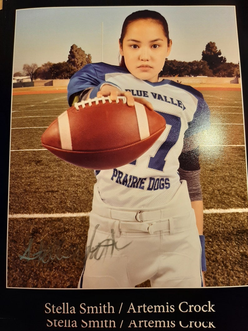 Może przedstawiać: Młoda kobieta w biało-niebieskim stroju piłkarskim z napisem "Blue Valley Prairie Dogs" trzyma brązowo-białą piłkę. Koszulka ma niebieski pasek na klatce piersiowej, a napis "Blue Valley" jest niebieski.