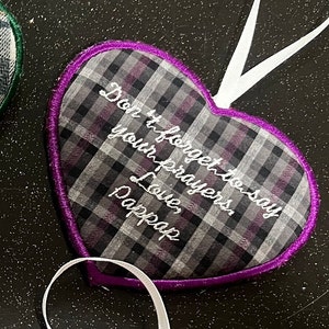 May include: Purple heart-shaped pillow with a gray and white plaid pattern. The pillow has a white ribbon attached and is embroidered with the text "Don't forget to say your prayers. Love, Pappap".