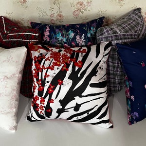 May include: Six decorative throw pillows with various patterns. The pillows are arranged in a row on a white surface. The pillows feature floral, zebra, and plaid patterns in shades of red, white, black, blue, and gray.