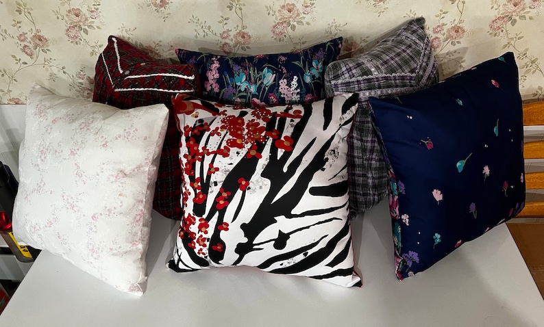 May include: Six decorative throw pillows with various patterns. The pillows are arranged in a row on a white surface. The pillows feature floral, zebra, and plaid patterns in shades of red, white, black, blue, and gray.