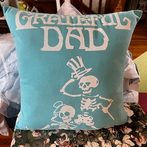 May include: A teal blue throw pillow with the text "Grateful Dad" in white lettering. The pillow features a white silhouette of two skeletons wearing top hats.