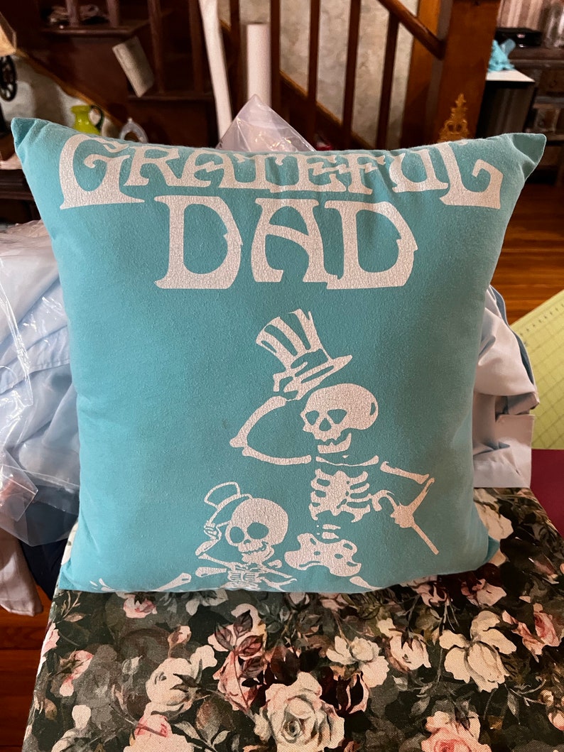 May include: A teal blue throw pillow with the text "Grateful Dad" in white lettering. The pillow features a white silhouette of two skeletons wearing top hats.