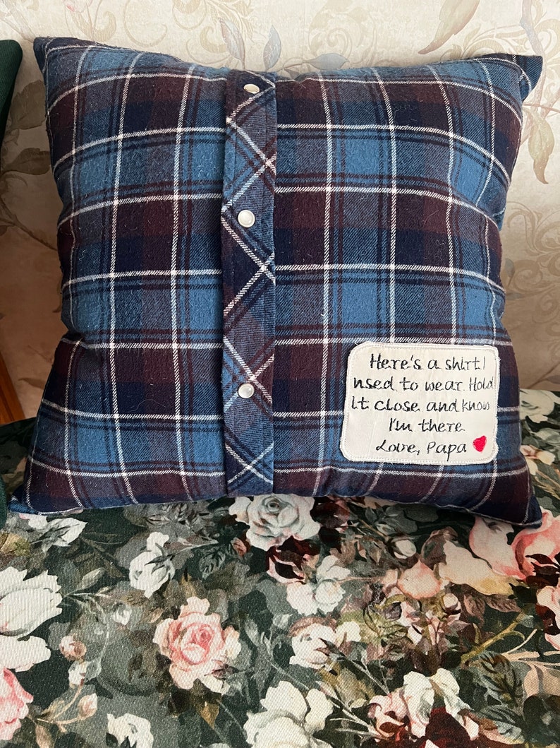 May include: A blue and brown plaid pillow with a white fabric patch sewn on. The patch has handwritten text that reads "Here's a shirt! used to wear. Hold it close and know I'm there Love, Papa" with a small red heart.