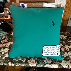 May include: A teal blue pillow with a white embroidered patch that says "Here's a shirt I used to wear. Hold it close and know I'm there. Love, Nathan" with a red heart. The pillow has a small black embroidered polo player logo in the upper right corner.