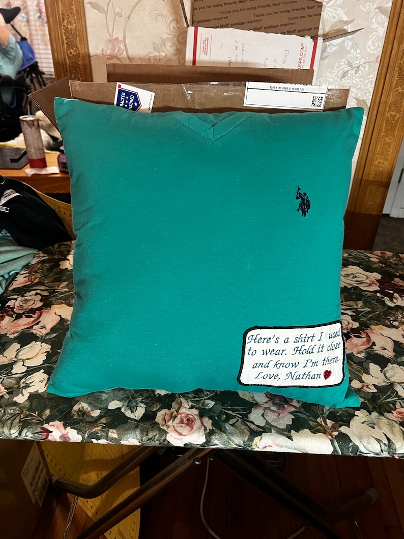 May include: A teal blue pillow with a white embroidered patch that says "Here's a shirt I used to wear. Hold it close and know I'm there. Love, Nathan" with a red heart. The pillow has a small black embroidered polo player logo in the upper right corner.