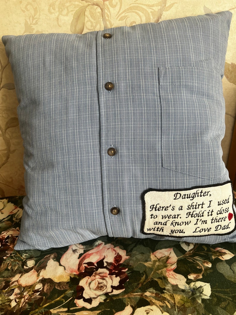 May include: A blue pillow with a pocket and buttons, made to look like a shirt. It has a white embroidered patch with the text "Daughter, Here's a shirt I used to wear. Hold it close and know I'm there with you, Love Dad."