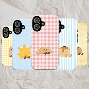 May include: A collection of phone cases with food-themed designs. The cases feature illustrations of a croissant, pancakes, and a sun, set against pastel gingham, grid, and striped backgrounds. The cases are in shades of yellow, blue, and pink.