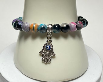 May include: A beaded bracelet with a hamsa hand charm featuring a blue evil eye. The bracelet is made with black, purple, orange, and blue agate beads.