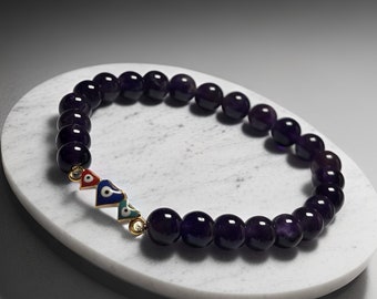 Puede incluir: Un brazalete de piedras preciosas de amatista púrpura con tres dijes dorados con ojos azules y rojos malvados. El brazalete está sobre una superficie de mármol blanco.