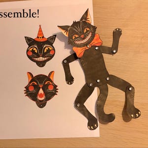 May include: A paper craft kit featuring a black cat figure with articulated limbs connected by silver rivets, an orange bow tie, and two interchangeable cat head cutouts, one with a witch hat, on a white sheet with the text Assemble!.