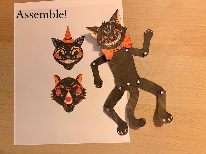 May include: A paper craft kit featuring a black cat figure with articulated limbs connected by silver rivets, an orange bow tie, and two interchangeable cat head cutouts, one with a witch hat, on a white sheet with the text Assemble!.