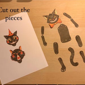 May include: A paper craft kit featuring a sheet with two black cat heads, one wearing an orange hat, and the text "Cut out the pieces". Beside the sheet are several detached black paper limbs, a torso, and a cat head with an orange bow tie.