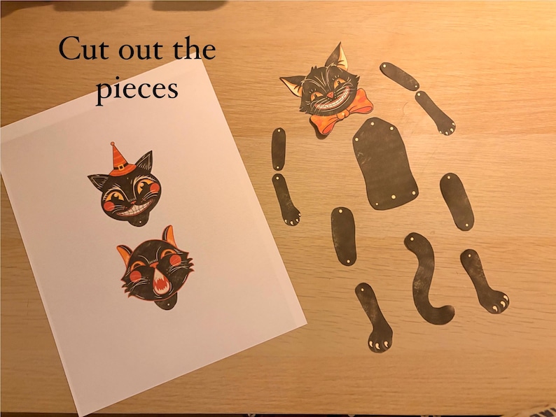 May include: A paper craft kit featuring a sheet with two black cat heads, one wearing an orange hat, and the text "Cut out the pieces". Beside the sheet are several detached black paper limbs, a torso, and a cat head with an orange bow tie.