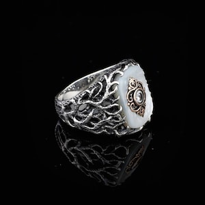 May include: A silver ring with a white mother-of-pearl stone set in a detailed, intricate design. The stone is surrounded by a gold-toned filigree pattern with a small clear stone in the center.