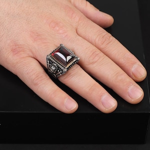 Può includere: Un anello d'argento con una grande pietra preziosa di colore rosso scuro incastonata in una cornice quadrata. L'anello presenta dettagli intricati e un design a forma di teschio.