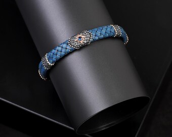May include: A blue braided leather bracelet with silver accents and a central star-shaped design. The bracelet features a woven texture and is adorned with small silver beads along the edges. The star has a blue gem in the center.