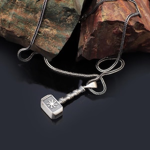 Puede incluir: Un collar plateado con un colgante en forma de martillo de Thor, Mjolnir. El colgante del martillo tiene un mango detallado y una cabeza cuadrada con un diseño en forma de estrella. La cadena es de estilo tejido. El fondo es oscuro.