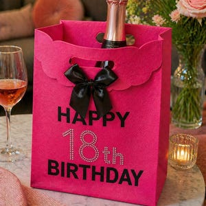 May include: A bright pink gift bag with a black bow and the text "HAPPY 18th BIRTHDAY" in black and rhinestones. A bottle of champagne is inside the bag. A wine glass and flowers are in the background.